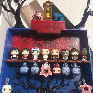 May include: A blue wooden display shelf featuring a red textured back panel, black tree branch accents, and a brown grandfather clock on top. The shelf holds eight small vinyl character figures on a ledge, with six more figures hanging below.