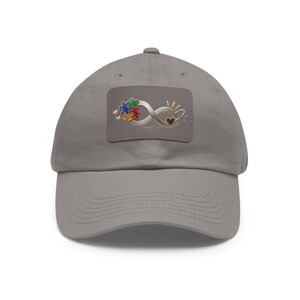 May include: A grey baseball cap featuring a rectangular patch on the front. The patch displays a metallic infinity symbol intertwined with a multi-colored puzzle piece design, small gold hearts, and a black heart, all set against a grey background.