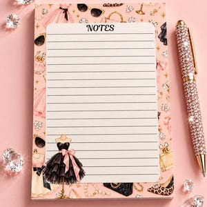 May include: A rectangular notepad with a pink background featuring illustrations of dresses, sunglasses, perfume bottles, and jewelry. The center has a white lined section with the word NOTES at the top. A pen covered in pink crystals sits to the right.