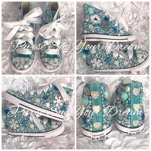 May include: A pair of turquoise high-top sneakers featuring white ribbon laces, adorned with various blue and white rhinestones, pearls, snowflake-shaped embellishments, and heart-shaped accents. The heels display the Converse All Star logo text.