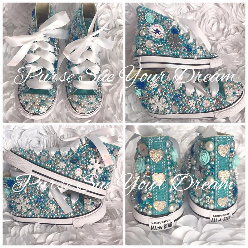 May include: A pair of turquoise high-top sneakers featuring white ribbon laces, adorned with various blue and white rhinestones, pearls, snowflake-shaped embellishments, and heart-shaped accents. The heels display the Converse All Star logo text.