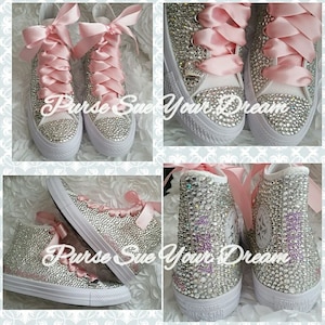 May include: High-top sneakers covered in clear crystals with wide pink satin ribbon laces. The shoes feature a white rubber toe cap and sole, with a silver heart-shaped charm engraved with the word Bride attached to the laces of one shoe.