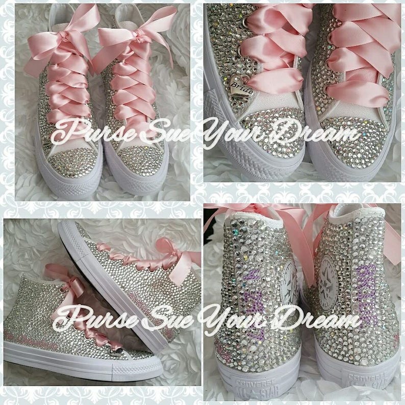 May include: High-top sneakers covered in clear crystals with wide pink satin ribbon laces. The shoes feature a white rubber toe cap and sole, with a silver heart-shaped charm engraved with the word Bride attached to the laces of one shoe.
