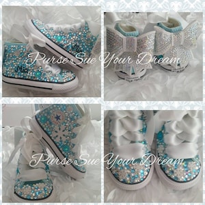 May include: Light blue high-top sneakers embellished with clear and blue rhinestones, white snowflake accents, and wide white ribbon laces. The heels feature large, sparkling white bows. The name Mia is written in black script on the side.