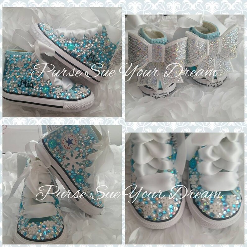 May include: Light blue high-top sneakers embellished with clear and blue rhinestones, white snowflake accents, and wide white ribbon laces. The heels feature large, sparkling white bows. The name Mia is written in black script on the side.