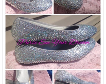 Op de afbeelding: Een paar zilveren ballerina's volledig bedekt met kleine, reflecterende, iriserende strass-steentjes. De schoenen hebben een ronde neus en een laag profiel. De tekst "Purse Sue Your Dream" is in roze schrift over de afbeeldingen heen geplaatst.