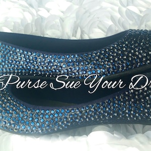 May include: A pair of dark blue ballet flats completely covered in small, sparkling blue rhinestones. The shoes feature a rounded toe and a low-profile silhouette. The text "Purse Sue Your Dream" is overlaid in white script across the center of the shoes.