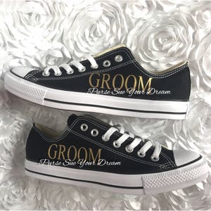 May include: A pair of black low-top sneakers with white laces and white rubber toe caps. The side of each shoe features the word GROOM in gold lettering, with the text Purse Sue Your Dream printed in white script underneath the gold text.