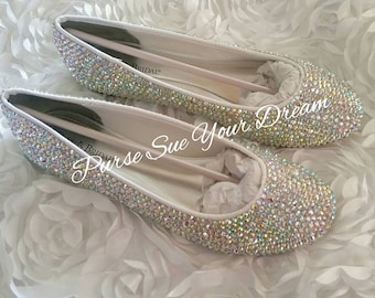 Op de afbeelding: Een paar witte ballerina's volledig bedekt met iriserende, lichtreflecterende kristallen. De binnenkant van de schoenen heeft een witte voering met de tekst "BRIDAL" zichtbaar op de binnenzool. De schoenen zijn gevuld met wit vloeipapier.