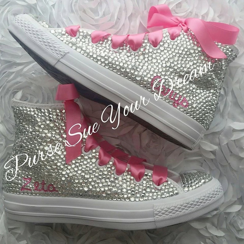 May include: High-top sneakers covered in clear crystals with bright pink satin ribbon laces and a pink bow at the top. The side of the shoe features the pink embroidered text Zeta, and the image includes the white cursive text Purse Sue Your Dream.