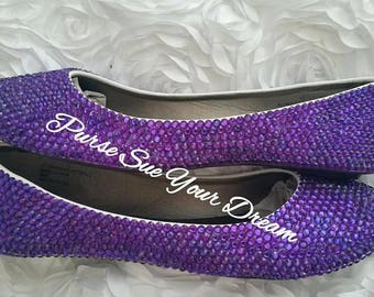 Op de afbeelding: Een paar paarse ballerina's volledig bedekt met kleine, reflecterende paarse strass-steentjes. De schoenen zijn voorzien van een witte binnenvoering en een witte tekst in script die "Purse Sue Your Dream" leest over het midden.
