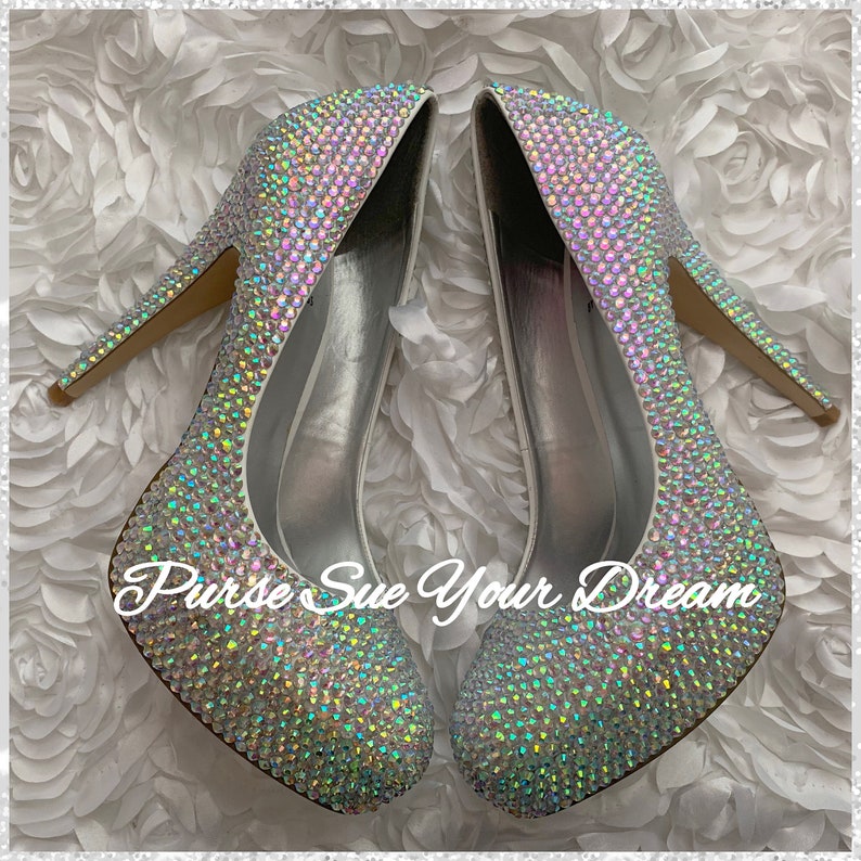 May include: A pair of high-heeled pumps completely covered in iridescent, light-reflecting crystals that shimmer with rainbow colors. The shoes feature a pointed toe and a slim heel. The text "Purse Sue Your Dream" is overlaid in white script.