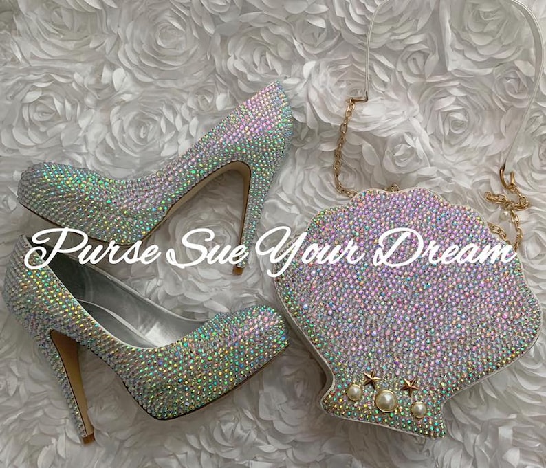 May include: A pair of high-heeled pumps and a shell-shaped clutch bag, both fully encrusted with iridescent crystals. The clutch features a gold chain strap and three pearl and star accents. The text "Purse Sue Your Dream" is overlaid on the image.