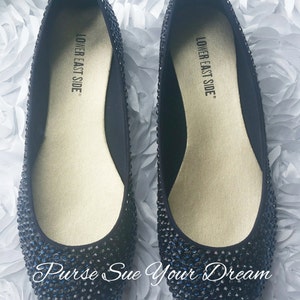 May include: A pair of black ballet flats covered in small, dark, reflective rhinestones. The interior insoles are gold-toned and feature the text "LOWER EAST SIDE" printed in black. The text "Purse Sue Your Dream" is visible at the bottom.