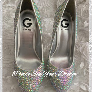 May include: A pair of G by Guess high-heeled pumps completely covered in iridescent, multi-colored rhinestones. The interior silver insoles feature the G by Guess logo. The text 'Purse Sue Your Dream' is overlaid in white script across the front.