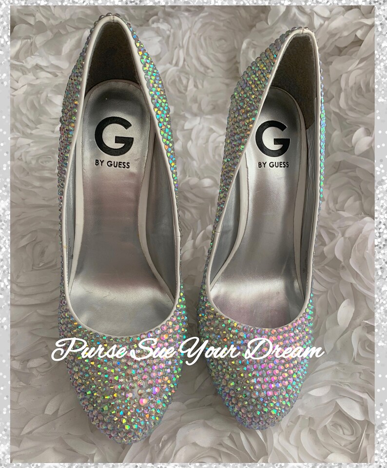 May include: A pair of G by Guess high-heeled pumps completely covered in iridescent, multi-colored rhinestones. The interior silver insoles feature the G by Guess logo. The text 'Purse Sue Your Dream' is overlaid in white script across the front.