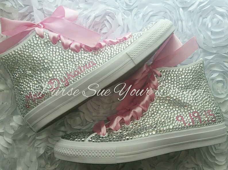 May include: A pair of high-top sneakers completely covered in clear rhinestones, featuring pink satin ribbon laces. The text Mrs. Dykatra is embroidered in pink on the side of one shoe, and the date 9.19.15 is embroidered on the other.