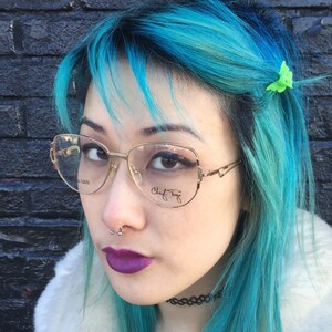Peut inclure: Une femme aux cheveux bleu vif et un piercing au nez porte des lunettes à monture dorée avec les mots "Claf  Tinge" sur la lentille droite. Elle porte une veste en fausse fourrure blanche et un collier ras du cou noir. Une pince à cheveux en plastique verte en forme de fleur est dans ses cheveux.