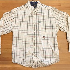 Könnte beinhalten: Weißes, langärmliges Hemd mit Knöpfen und einem schwarzen und roten Karomuster. Das Hemd hat ein kleines gesticktes Logo auf der linken Brust.