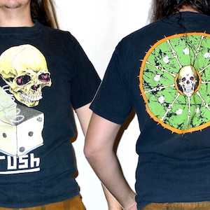 May include: Black t-shirt with a graphic of a skull in a box with a spring and dice. The text "Rush" is printed below the graphic. The back of the shirt has a green and orange graphic of a skull with bones and dice.