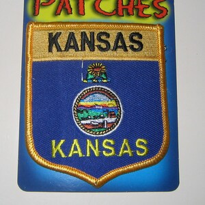 May include: A blue and gold embroidered patch with the state seal of Kansas and the word "Kansas" written twice. The patch is on a blue and white card with the word "Patches" written in red.