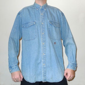 Könnte beinhalten: Ein hellblaues Jeanshemd mit langen Ärmeln, Knopfleiste und zwei Brusttaschen. Das Hemd hat einen kleinen Logo-Patch auf der linken Brust.