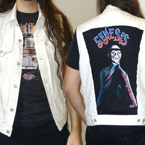 Puede incluir: Un chaleco vaquero blanco con una camiseta negra debajo. El chaleco tiene un gran parche negro en la espalda con la palabra "Genesis" en letras rojas, azules y blancas. El parche presenta una imagen en blanco y negro de una persona con una máscara y una capa.