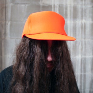 May include: A person with long brown hair is wearing a bright orange baseball cap. The cap has a curved brim and a mesh back.