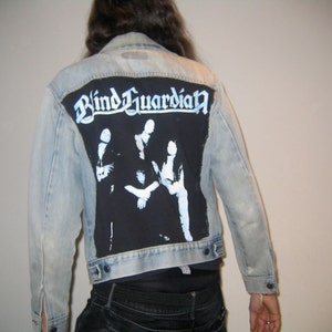 Pode incluir: Uma jaqueta de ganga azul claro com um remendo preto nas costas. O remendo apresenta um logotipo de banda que diz "Blind Guardian" em letras brancas e uma imagem em preto e branco de uma banda.