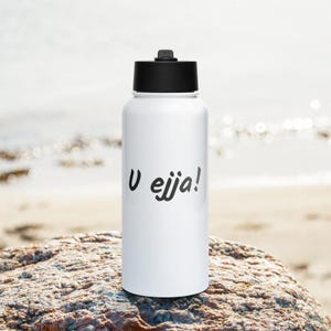 Könnte beinhalten: Eine weiße Wasserflasche mit einem schwarzen Schraubverschluss. Auf der Vorderseite der Flasche befindet sich der schwarze Schriftzug U ejja! in einer stilisierten Schreibschrift. Die Flasche hat einen glatten, zylindrischen Körper und einen flachen Boden.