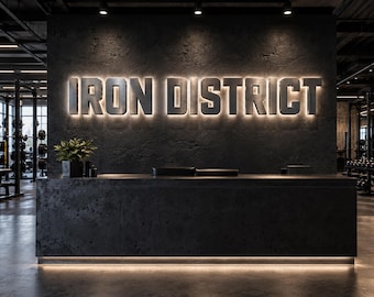 May include: A dark gray reception desk sits in front of a textured black wall featuring the illuminated silver text IRON DISTRICT. The gym interior includes metal weight racks and equipment visible in the background under soft lighting.