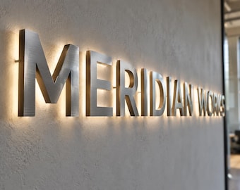 May include: A set of brushed silver metal 3D letters spelling out MERIDIAN WORKS mounted on a textured wall. The letters feature a back-lit design that casts a warm, soft glow onto the surface behind each individual character.