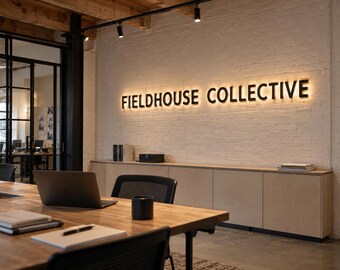 May include: A white brick wall features the black, backlit text FIELDHOUSE COLLECTIVE. Below the sign is a long, light-colored wooden cabinet. In the foreground, a wooden desk holds a laptop, a black cup, and notebooks.