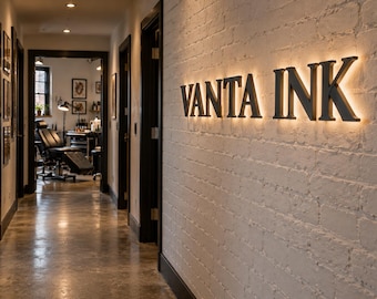 May include: A white painted brick wall features the black backlit lettering VANTA INK. The wall is located in a hallway with a polished concrete floor leading to a room containing a black tattoo chair and professional equipment.