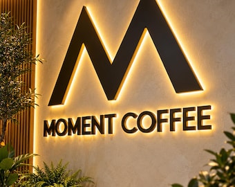 May include: A large, black, three-dimensional letter M mounted on a textured wall above the text MOMENT COFFEE. The signage features warm, backlit LED lighting that creates a glowing halo effect around the dark, bold lettering.