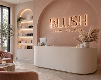 May include: A reception area featuring a curved white desk and a wall with a large arched niche. The wall displays the illuminated text BLUSH BROW STUDIO in metallic lettering. Shelving units with integrated lighting hold various products.