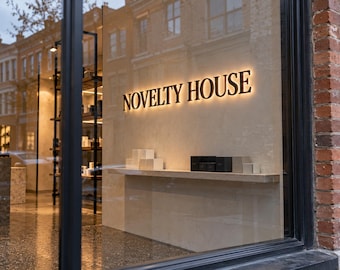May include: A storefront window display featuring the text NOVELTY HOUSE in backlit, dark, dimensional lettering mounted on a light-colored wall. A horizontal shelf holds several small, square, neutral-toned product boxes.