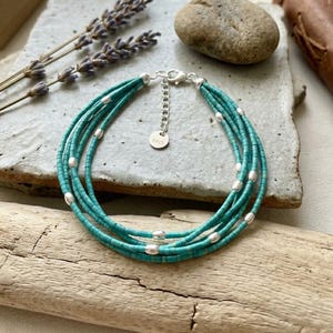 Op de afbeelding: Een armband met meerdere strengen, bestaande uit kleine turquoise kralen afgewisseld met gepolijste zilverkleurige metalen tussenstukjes. De armband is voorzien van een zilverkleurige verlengketting en een rond plaatje met de inscriptie 925.