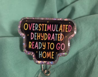 Puede incluir: Un portacredenciales negro con borde brillante y texto colorido que dice: OVERSTIMULATED DEHYDRATED READY TO GO HOME. El portacredenciales cuenta con una correa de plástico transparente con un broche de metal para sujetar tarjetas de identificación.