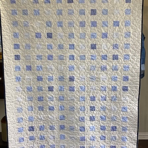 May include: A white quilt with a blue and white checkered pattern. The quilt is made up of many small squares, each with a different pattern. The quilt is hanging on a stand.