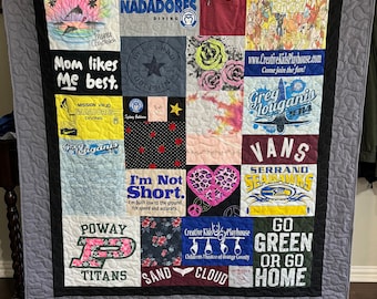 May include: A quilt made from a variety of t-shirts, featuring logos, text, and images. The quilt is grey with black borders and is displayed on a wooden stand. Some of the text on the quilt includes "Mom likes me best", "I'm not short", "Go Green or Go Home", and "Creative Kids Playhouse".