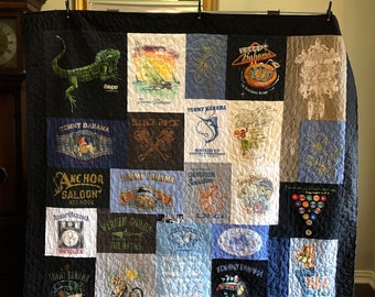 May include: A quilt made from a variety of t-shirts, featuring a black background and a patchwork design. The t-shirts feature various designs, including a lizard, a sailboat, a palm tree, and a bar. The quilt is signed "Larry Torrence".