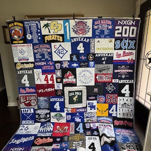 以下が含まれることがあります： 様々なスポーツチームのロゴと数字をパッチワークデザインにした手作りのキルト。キルトは、青、赤、白など様々な色を取り入れており、「Pirates」、「Saints」、「All Stars」などの文字が使用されています。