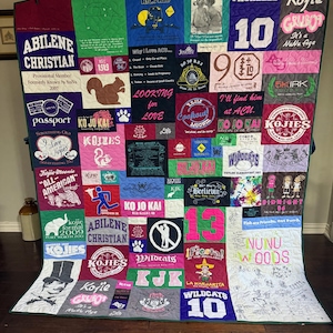 Pode incluir: Uma colcha de memórias feita de vários painéis de t-shirts coloridas com diversos estampados gráficos, texto e logótipos. O design de patchwork inclui numerosos slogans, números e ilustrações impressas cosidos em grelha.