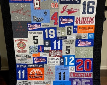 May include: A patchwork quilt made from repurposed t-shirts, displaying a variety of logos and text in shades of blue, red, and grey. The quilt features the words "Christian," "German," and "Marco Island." It is finished with a black border.