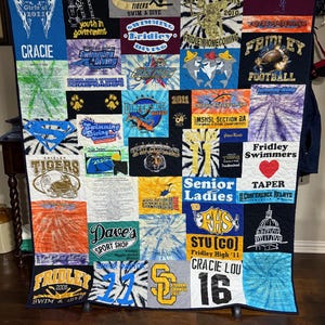 Pode incluir: Uma grande colcha retangular construída a partir de vários painéis de t-shirts coloridas com diversos estampados gráficos, logótipos escolares, emblemas de equipas desportivas e texto como Fridley Tigers, Senior Ladies e Gracie Lou.