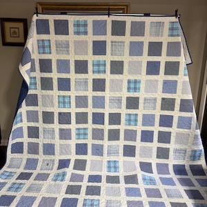 Könnte beinhalten: Eine große, handgefertigte Steppdecke mit einem Gittermuster aus blauen Quadraten auf weißem Hintergrund. Die Quadrate weisen verschiedene blaue Muster auf, darunter Karos und unifarbene Farben. Die Steppdecke hat einen blauen Rand.
