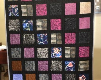 May include: A quilt made of various fabrics in shades of purple, pink, gray, and brown. The quilt is made up of 36 squares, each with a different pattern. The quilt is hanging on a wall.