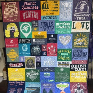Op de afbeelding: Een veelkleurige t-shirtquilt bestaande uit een raster van diverse bedrukte stoffen vierkanten. De quilt toont talloze grafische ontwerpen, logo's en inspirerende teksten met betrekking tot de Woodlands Christian Academy, inclusief verwijzingen naar krijgers, geloof en schoolevenementen.