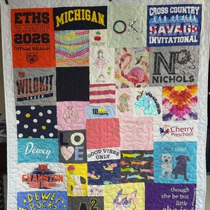 Peut inclure: Une courtepointe souvenir composée de divers carrés de tissu présentant des imprimés graphiques variés, incluant des textes comme ETHS Class of 2026, Michigan, Cross Country Savage Invitational, Nichols, Cherry Preschool et la citation though she be but little she is fierce.
